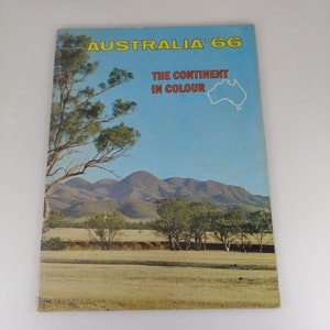 Può includere: Un libro di viaggio vintage intitolato "AUSTRALIA '66" con una scena paesaggistica sulla copertina. La copertina presenta un cielo blu, montagne, alberi e il testo "THE CONTINENT IN COLOUR". Il libro è leggermente usurato.