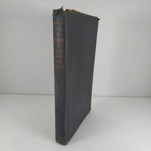 May include: A vintage hardcover book with a dark blue cover and gold lettering on the spine. The book's title and author are visible. The top edge of the book shows signs of wear and tear, suggesting its age. The book is standing upright.