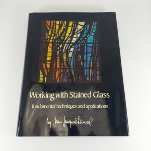 May include: A black book titled "Working with Stained Glass" with a colourful stained glass window design on the cover. The book's subtitle reads "fundamental techniques and applications" and is signed by Jean Jacques Duval.