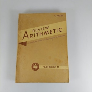 Peut inclure: Un manuel scolaire brun intitulé "Review Arithmetic" avec le sous-titre "Decimals, Per Cents, and Applications of Arithmetic". Le livre est intitulé "Textbook II" et porte le logo USAFI sur la couverture.