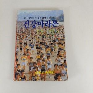 Puede incluir: Una portada de libro vintage con una foto de un gran grupo de personas corriendo en un maratón. El título del libro es "건강마라톤" en coreano, que se traduce como "Maratón de la salud". El texto "뛰는 것보다 더 좋은 補藥은 없다!" también es visible en la portada, que se traduce como "¡No hay mejor medicina que correr!"