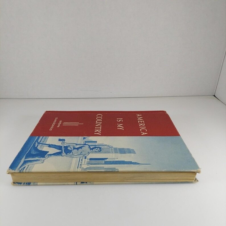 Puede incluir: Un libro rojo y azul con el t&iacute;tulo "America Is My Country" en la portada. La portada presenta una ilustraci&oacute;n azul de un hombre sosteniendo un martillo frente a un horizonte urbano.
