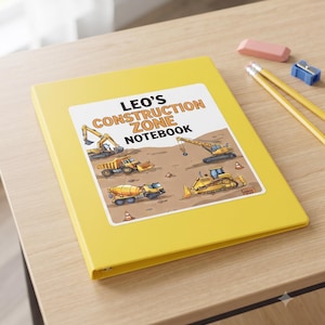 Op de afbeelding: Een felgele ringband met drie ringen en een centraal label met de tekst LEO'S CONSTRUCTION ZONE NOTEBOOK. Het label toont cartoonillustraties van gele bouwvoertuigen, waaronder een graafmachine, een kraan en een betonmixer.