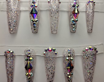 May include: Set of ten long, clear, coffin-shaped false nails with iridescent glitter and rhinestones. The nails have a silver glitter base and are decorated with iridescent rhinestones in a variety of shapes and sizes.