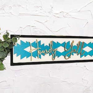 May include: A wooden sign with a black frame and a turquoise and white geometric pattern. The sign says "Howdy Y'all" in gold lettering.