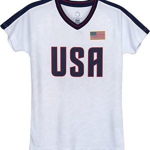 Puede incluir: Camiseta blanca de manga corta con ribetes azul marino y rojo. La camiseta tiene cuello en V y presenta la palabra "USA" en azul marino con contorno rojo. Una peque&ntilde;a bandera estadounidense se encuentra en el lado derecho del pecho.