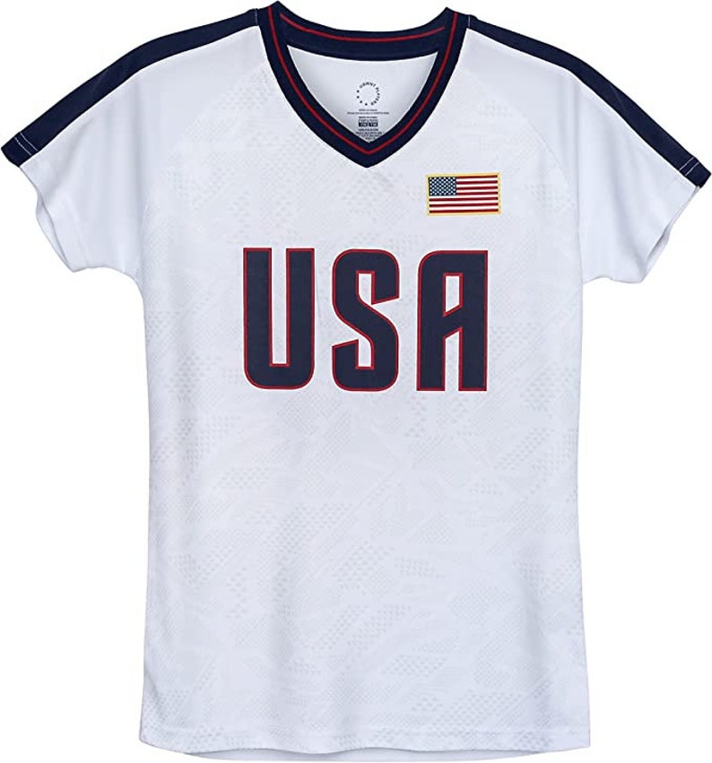 Puede incluir: Camiseta blanca de manga corta con ribetes azul marino y rojo. La camiseta tiene cuello en V y presenta la palabra "USA" en azul marino con contorno rojo. Una peque&ntilde;a bandera estadounidense se encuentra en el lado derecho del pecho.