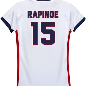 May include: White soccer jersey with red and blue trim. The back of the jersey has the name "RAPINOE" and the number "15" printed in blue with a red outline.