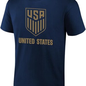 Pu&ograve; includere: Una maglietta blu navy con uno scudo dorato che presenta le lettere "USA" e strisce verticali. Le parole "UNITED STATES" sono stampate sotto lo scudo.