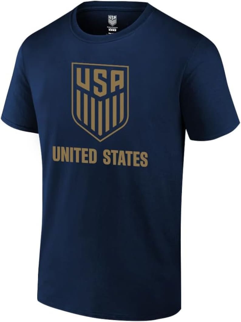 Pu&ograve; includere: Una maglietta blu navy con uno scudo dorato che presenta le lettere "USA" e strisce verticali. Le parole "UNITED STATES" sono stampate sotto lo scudo.