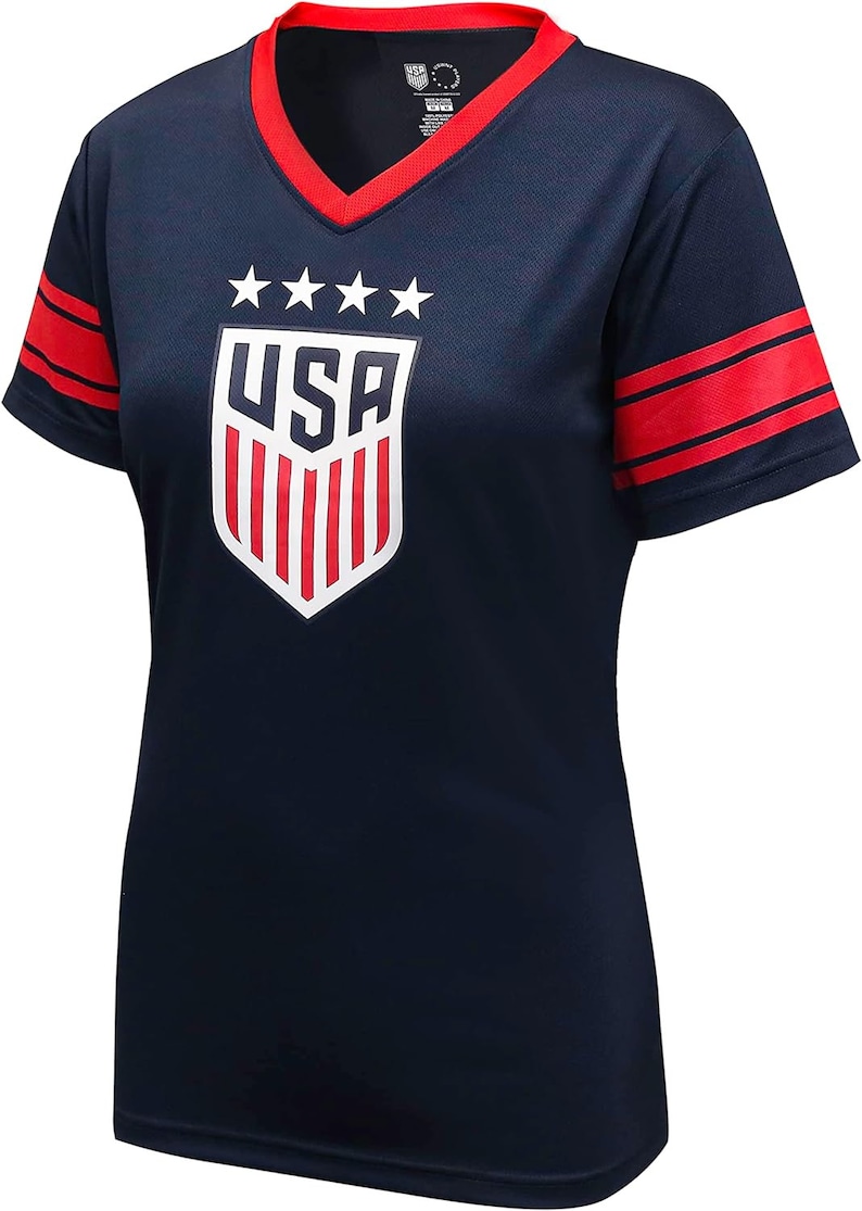 May include: A navy blue short-sleeved t-shirt with red and white stripes on the sleeves. The shirt features a white shield with red stripes and the word "USA" in white letters. There are five white stars above the shield.