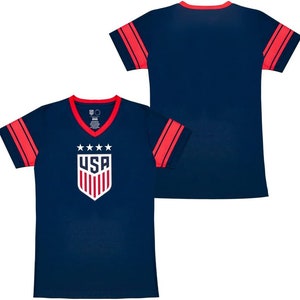 May include: A navy blue t-shirt with red and white stripes on the sleeves. The front of the shirt features a white "USA" logo with five stars and a shield design.