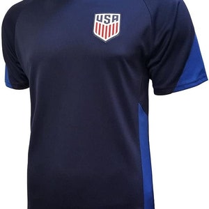 May include: A navy blue soccer jersey with a red, white, and blue USA crest on the front. The jersey has blue accents on the sleeves and sides.