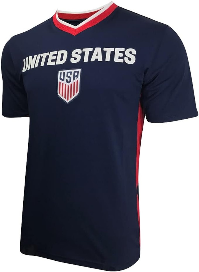 May include: A navy blue football shirt with red and white trim. The shirt has the words "UNITED STATES" printed on the front, along with a shield with the letters "USA" inside.