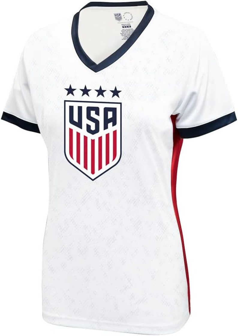 K&ouml;nnte beinhalten: Wei&szlig;es Fu&szlig;balltrikot mit einem blauen und roten USA-Wappen und f&uuml;nf Sternen &uuml;ber dem Wappen. Das Trikot hat einen dunkelblauen Kragen und dunkelblaue Einfassungen an den &Auml;rmeln. Das Trikot hat einen roten Streifen auf der rechten Seite.