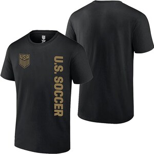 K&ouml;nnte beinhalten: Schwarzes T-Shirt mit einem goldenen "U.S. SOCCER"-Motiv auf der rechten Seite und einem goldenen "USA"-Logo auf der linken Seite.