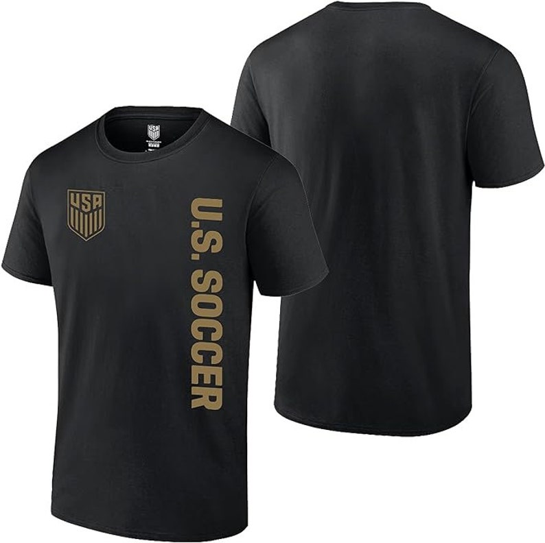 K&ouml;nnte beinhalten: Schwarzes T-Shirt mit einem goldenen "U.S. SOCCER"-Motiv auf der rechten Seite und einem goldenen "USA"-Logo auf der linken Seite.