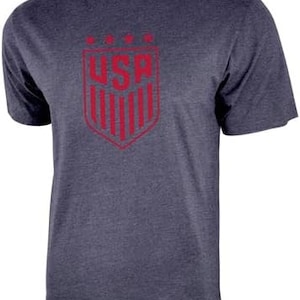May include: A navy blue t-shirt with a red USA crest graphic. The crest features a shield with vertical stripes and the letters USA in a bold font.