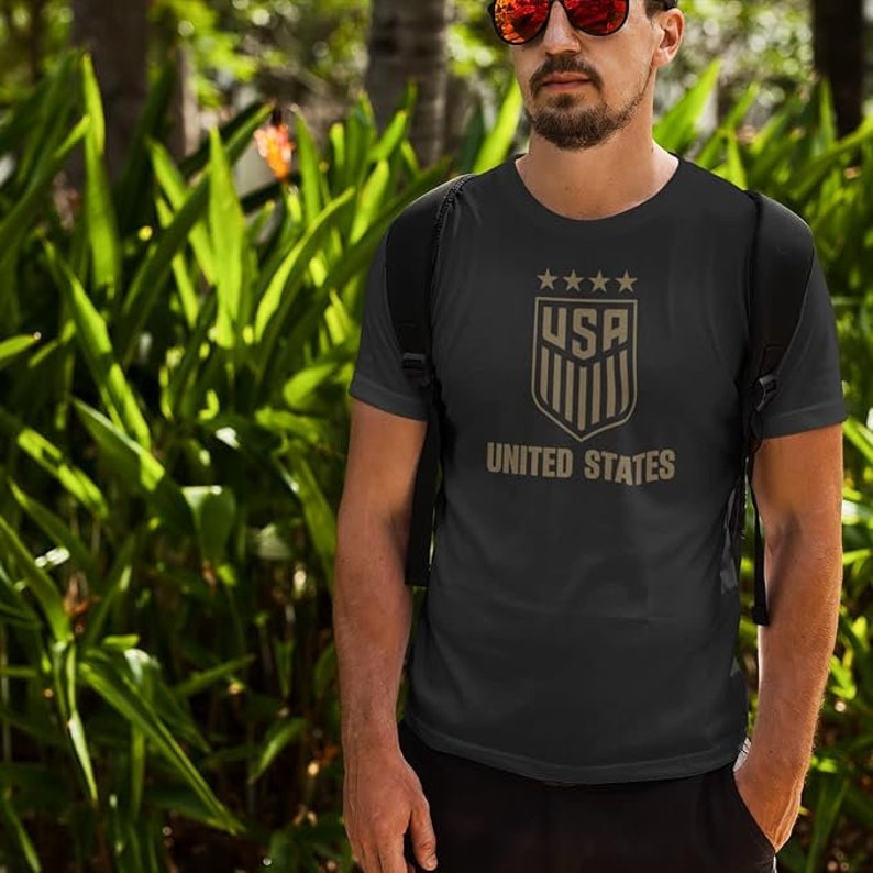 May include: A black t-shirt with a gold graphic of the United States soccer team logo. The logo features a shield with the letters "USA" and five stars above the shield. The text "UNITED STATES" is below the shield.
