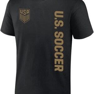 K&ouml;nnte beinhalten: Schwarzes T-Shirt mit einem goldenen U.S. Soccer-Logo auf der linken Brust und "U.S. SOCCER" vertikal auf der rechten Seite gedruckt.