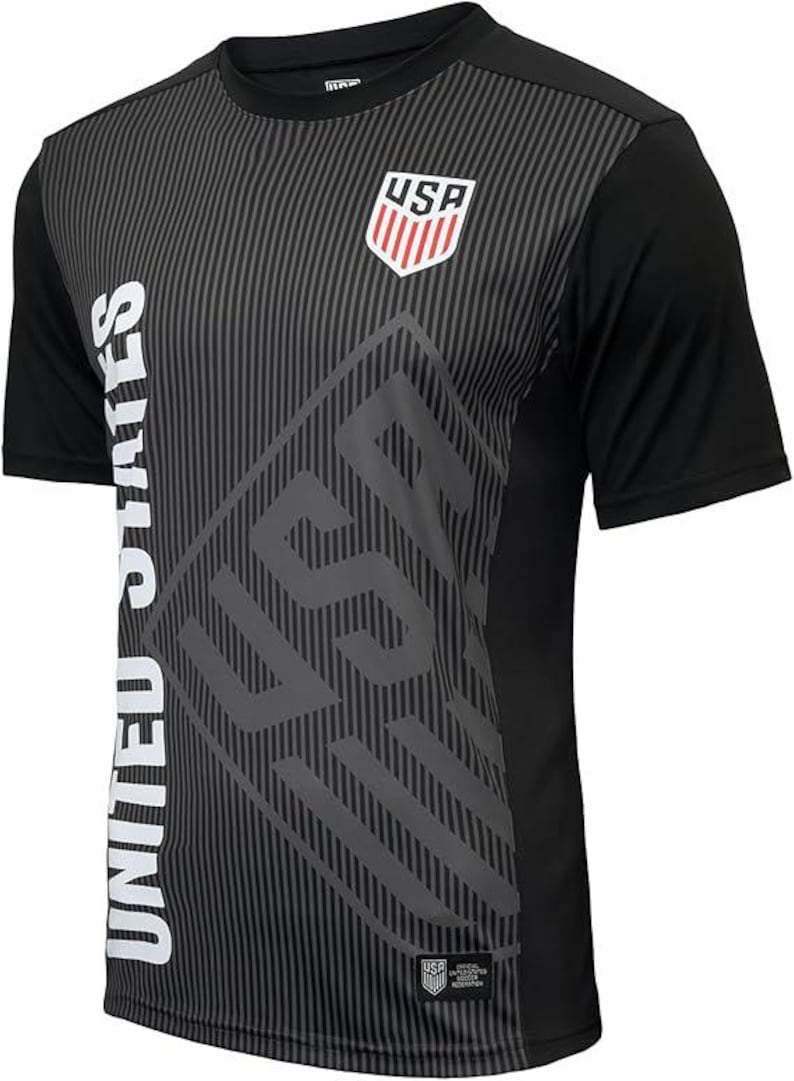 Puede incluir: Camiseta de f&uacute;tbol negra con un estampado gr&aacute;fico blanco de "United States" y "USA". La camiseta tiene un patr&oacute;n de rayas negras y grises.