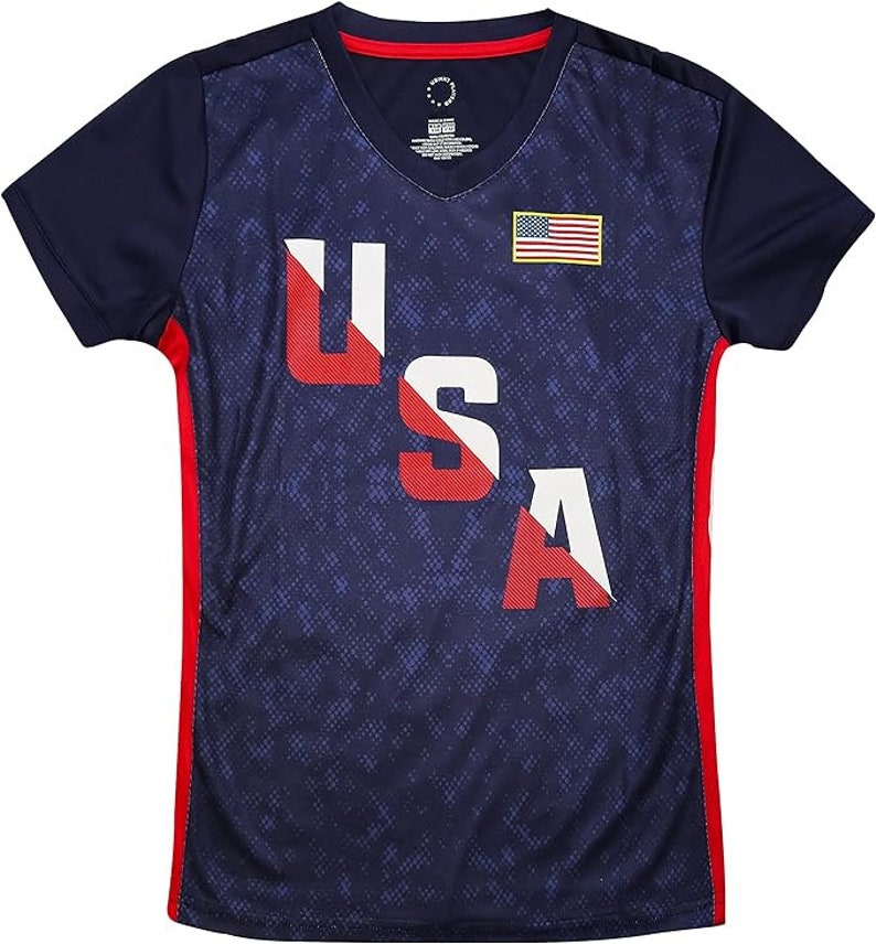 Pu&ograve; includere: Una maglietta blu navy con una grafica USA in rosso, bianco e blu e una piccola bandiera americana sul petto. La maglietta ha pannelli laterali rossi e un collo a V.