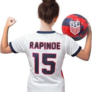May include: A white soccer jersey with blue trim and the name "RAPINOE" and the number "15" printed on the back. The jersey is being held by a person holding a red, white, and blue soccer ball with the word "USA" printed on it.