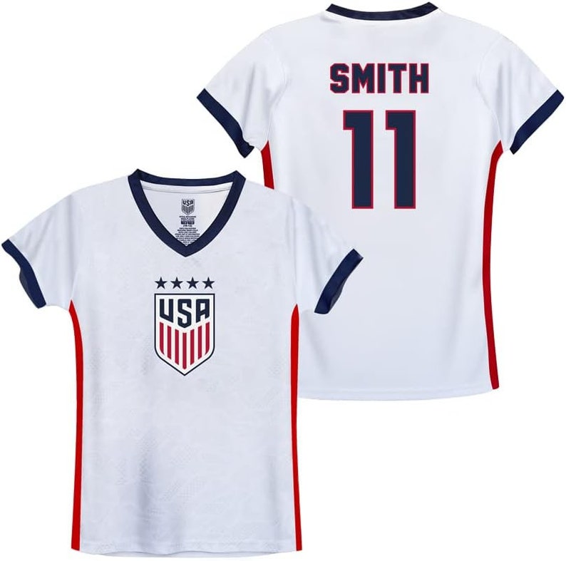 Puede incluir: Camiseta de f&uacute;tbol blanca con ribetes rojos y azules. La camiseta tiene el logotipo de EE. UU. en la parte delantera y el nombre "SMITH" y el n&uacute;mero "11" en la parte posterior.