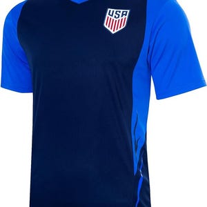 Pode incluir: Uma camisa de futebol azul marinho com um design de chama azul e branca no lado direito. A camisa tem um decote em V e apresenta um logotipo dos EUA vermelho, branco e azul no peito.