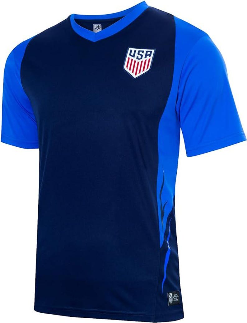 Pode incluir: Uma camisa de futebol azul marinho com um design de chama azul e branca no lado direito. A camisa tem um decote em V e apresenta um logotipo dos EUA vermelho, branco e azul no peito.