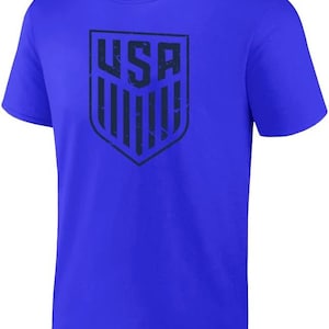 May include: A royal blue t-shirt featuring a distressed navy blue graphic of the USA shield logo.