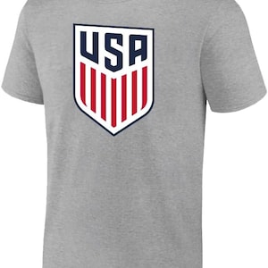 May include: A gray t-shirt with a navy blue and red USA logo on the front. The logo is a shield with the word "USA" in white letters and red and white stripes.