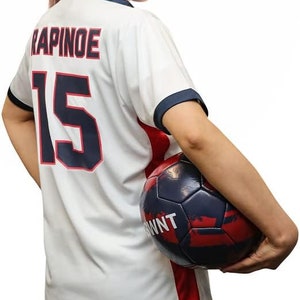 May include: A white soccer jersey with the name "RAPINOE" and the number "15" on the back. The jersey has red and blue trim. A person is holding a soccer ball with a red, white, and blue design.