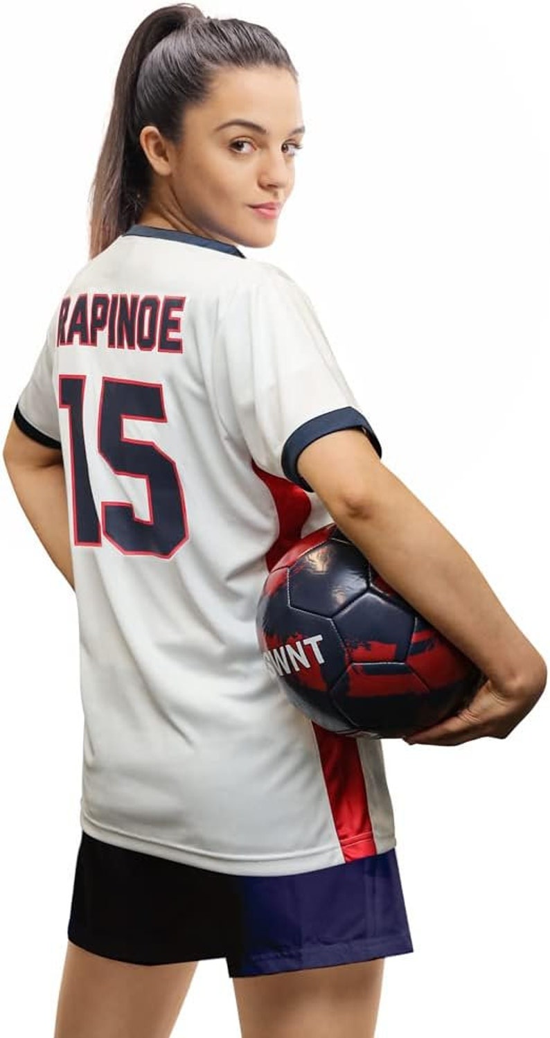 May include: A white soccer jersey with the name "RAPINOE" and the number "15" on the back. The jersey has red and blue trim. A person is holding a soccer ball with a red, white, and blue design.