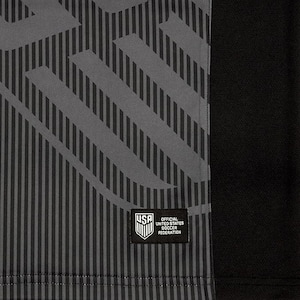 Puede incluir: Camiseta de f&uacute;tbol negra y gris con un patr&oacute;n geom&eacute;trico y un parche blanco con el texto "USA Official United States Soccer Federation".