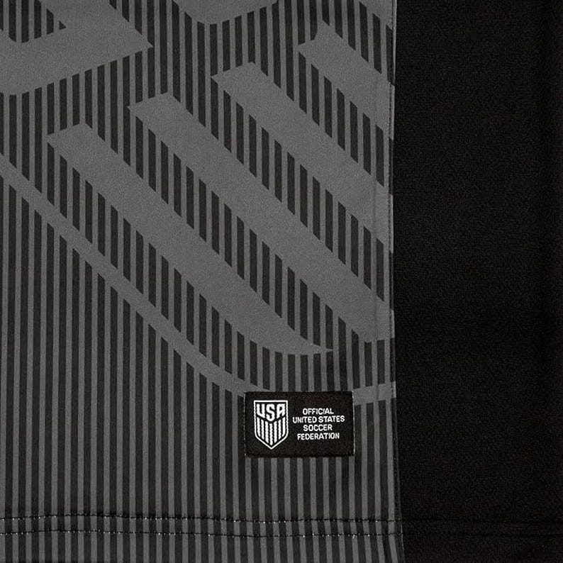 Puede incluir: Camiseta de f&uacute;tbol negra y gris con un patr&oacute;n geom&eacute;trico y un parche blanco con el texto "USA Official United States Soccer Federation".