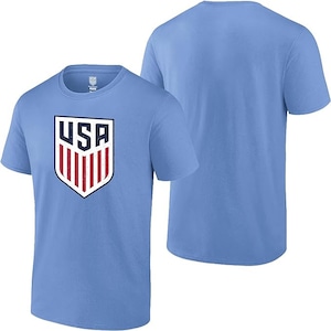 Pode incluir: Camiseta azul clara com um design de escudo com as letras "USA" em branco e azul marinho, e listras vermelhas e brancas. A camiseta tem gola redonda e mangas curtas. Outra camiseta &eacute; vis&iacute;vel no fundo.