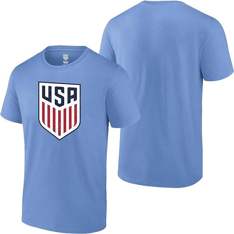 Pode incluir: Camiseta azul clara com um design de escudo com as letras "USA" em branco e azul marinho, e listras vermelhas e brancas. A camiseta tem gola redonda e mangas curtas. Outra camiseta &eacute; vis&iacute;vel no fundo.