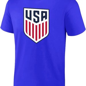 Pode incluir: Uma camiseta azul royal com um escudo branco e vermelho com a palavra "USA" em letras azul marinho.