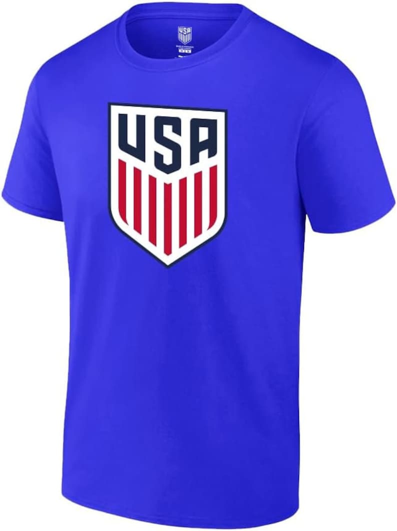 Pode incluir: Uma camiseta azul royal com um escudo branco e vermelho com a palavra "USA" em letras azul marinho.