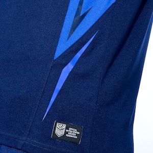 Pode incluir: Uma camisa de futebol azul marinho com um design de raio e um patch que diz "Official United States Soccer Federation".