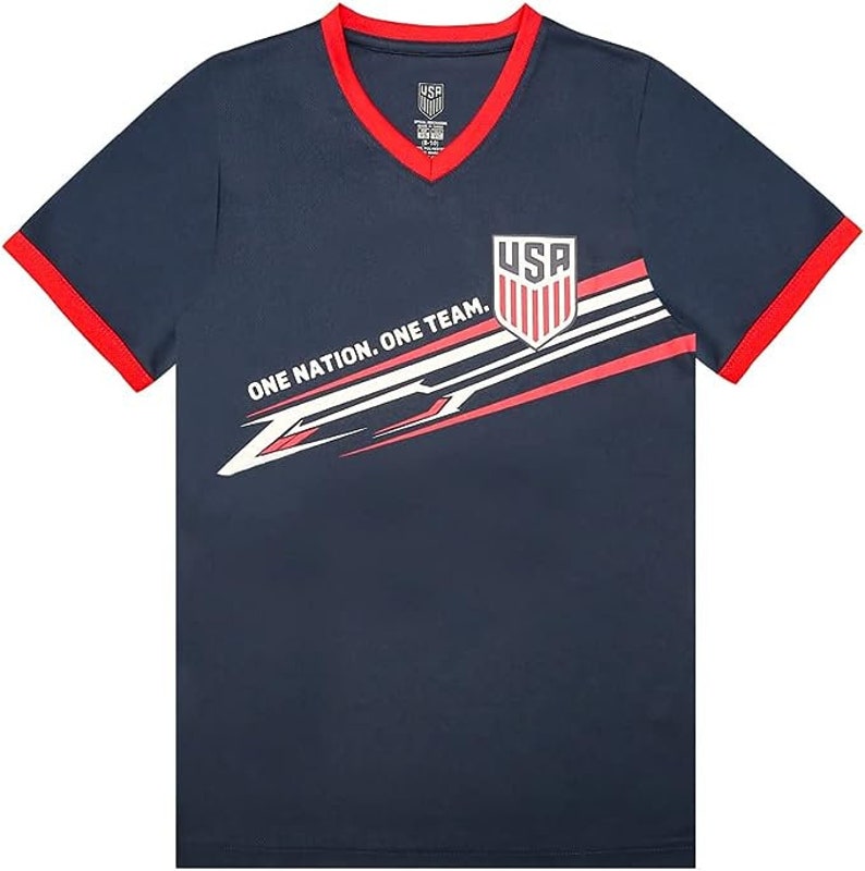 May include: A navy blue t-shirt with red and white accents. The shirt features a graphic of a shield with the word "USA" and the text "One Nation. One Team."