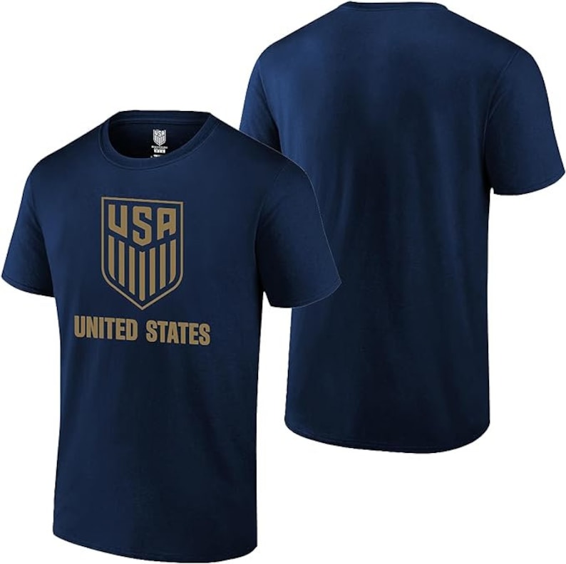 Pu&ograve; includere: Una t-shirt blu navy con uno stemma dorato che presenta le lettere "USA" e le parole "UNITED STATES".