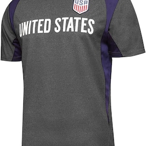 May include: A heather grey t-shirt with purple accents on the sleeves. The shirt has a white graphic of the United States flag and the text "UNITED STATES".
