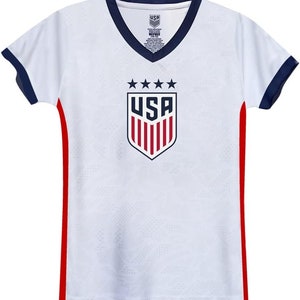 Puede incluir: Camiseta de f&uacute;tbol blanca con ribetes rojos y azules. La camiseta tiene un gran escudo de los Estados Unidos con cinco estrellas encima.