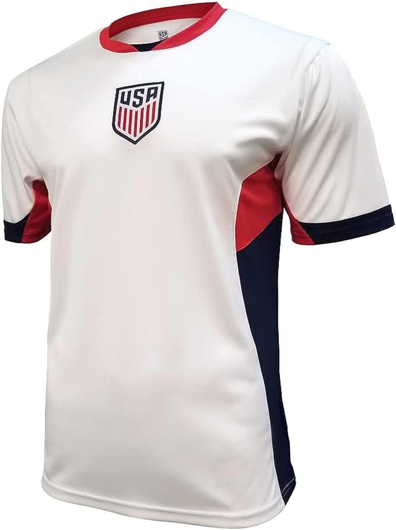 Op de afbeelding: Wit voetbaltenue met een rood, wit en blauw USA-wapen op de voorkant. Het tenue heeft rode en donkerblauwe accenten op de mouwen en zijkanten.