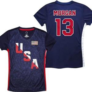 Pu&ograve; includere: Due maglie da calcio blu navy. Una maglia ha il testo "USA" in rosso, bianco e blu con una piccola bandiera americana sul petto. L'altra maglia ha il testo "MORGAN" e il numero "13" in rosso.