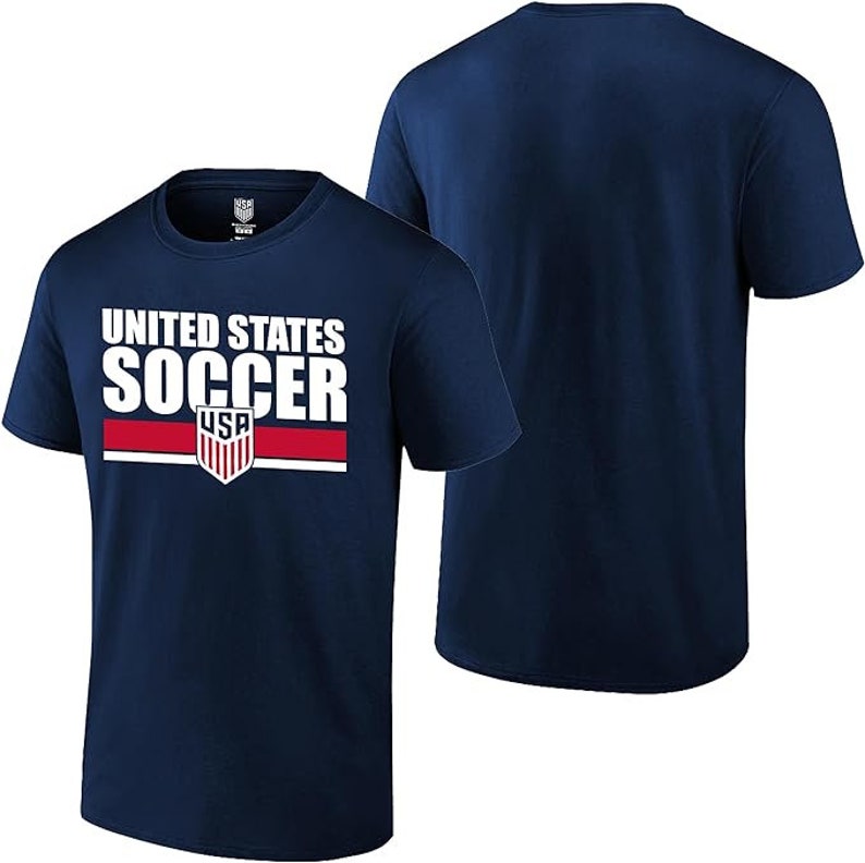 May include: Navy blue t-shirt featuring the text "UNITED STATES SOCCER" in white. A red and white stripe is below the word "SOCCER" with a USA shield logo. The shirt is short-sleeved.