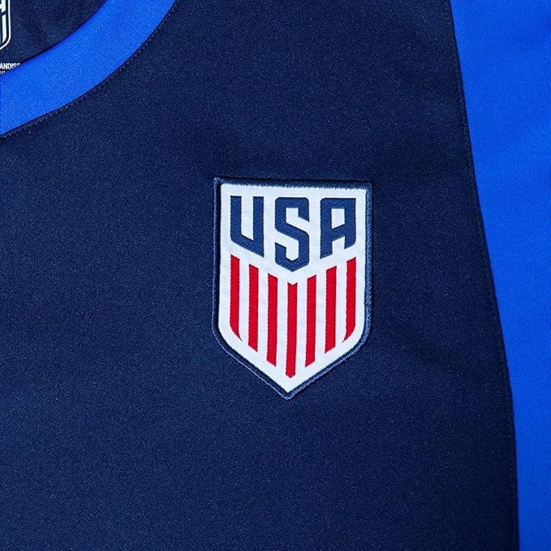 Pode incluir: Uma camisa de futebol azul marinho com um bras&atilde;o dos EUA branco e vermelho na frente. A camisa tem detalhes azuis nas mangas.