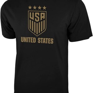 May include: Black t-shirt with a gold graphic of the United States soccer team logo. The logo features five stars above the letters "USA" and a shield with vertical stripes. The text "UNITED STATES" is printed below the logo.